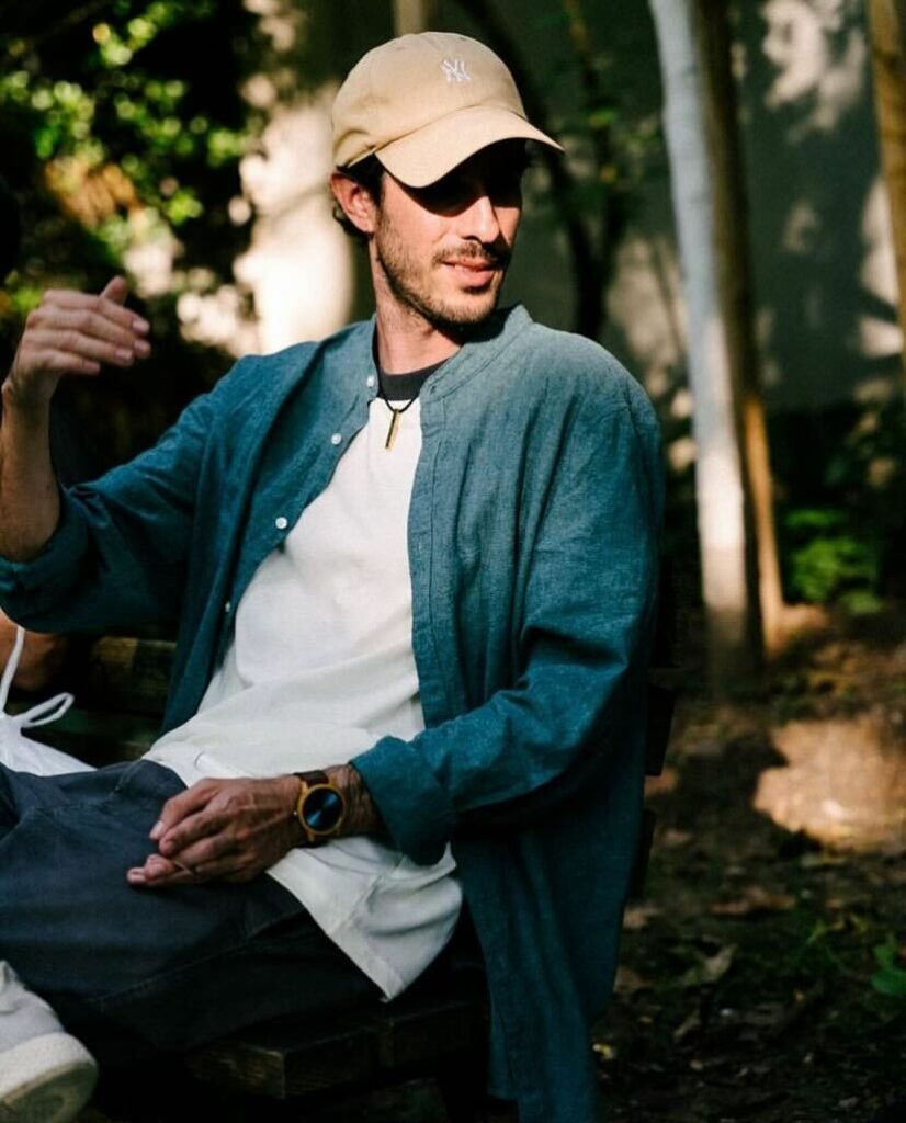 Nadav Elovic, a content creator, sitting outdoors, wearing a beige cap and a blue shirt, gesturing while engaged in conversation. Perfect for stock footage and video editing projects focused on casual and natural settings