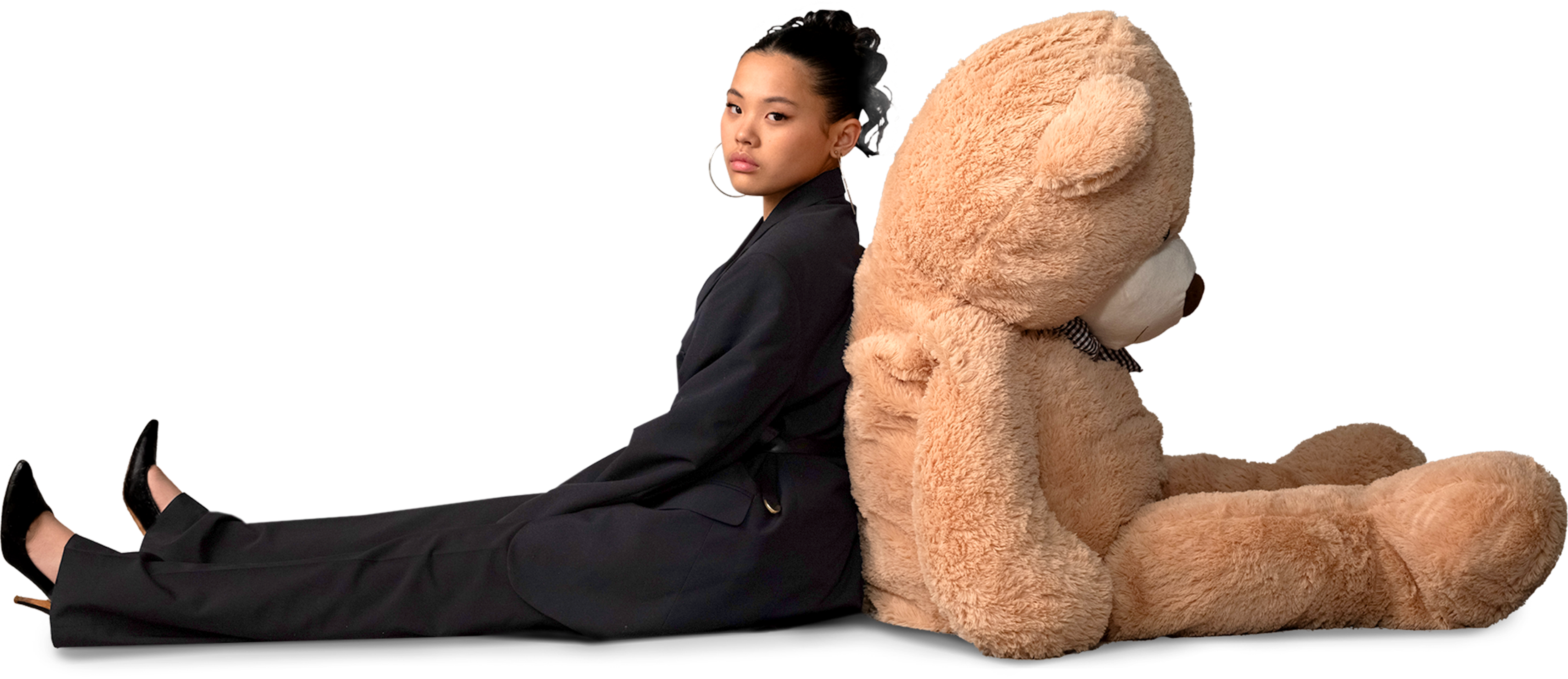 A content creator dressed in black leans against a large teddy bear, both seated on the floor, creating a whimsical and contrasting visual